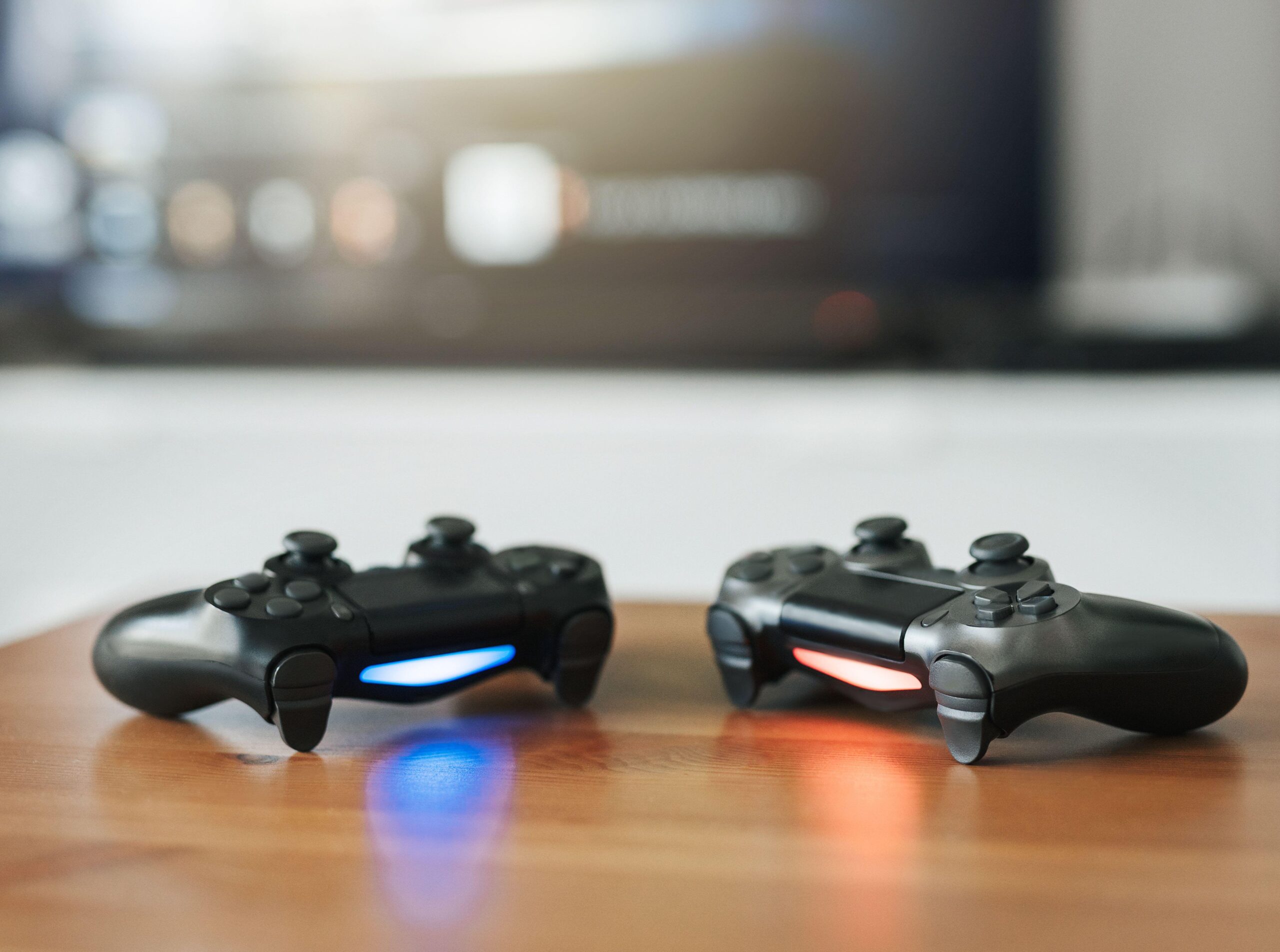 current console generation, Playstation 5 controllers on a wooden table with a game on the monitor behind them